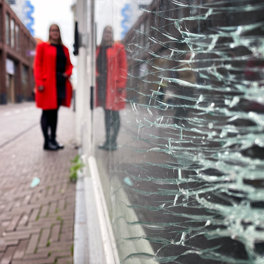 Lelijk is ook mooi: een detail van een kapotte etalageruit in Den Haag met daarin de reflectie van een dame met een rode jas
