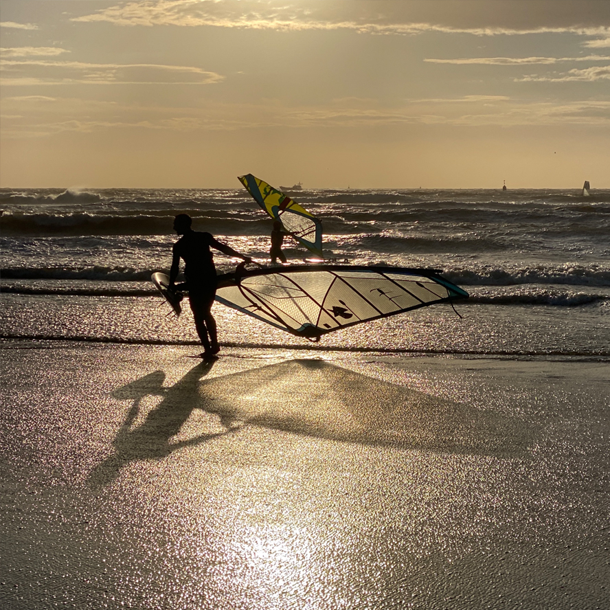 Surfer in de branding met zeil en plank in warm, geel tegenlicht tijdens het gouden uur